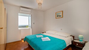 A bedroom with a bed, a window, a chandelier, a painting, and a nightstand with a lamp.