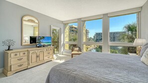Un dormitorio con una cama grande, un aparador con cajones, un televisor, un espejo y ventanas amplias con vistas a edificios y árboles.