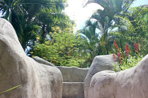 A stone pathway flanked by large boulders, leading through a lush garden with various trees and flowering plants.