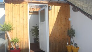 Fachada de madera con una puerta que da a un espacio interior. Hay plantas en macetas en el suelo.