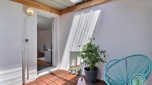 Un balcón con suelo de madera, una planta en maceta, una silla azul y una cortina blanca.