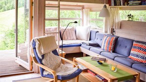 Ein gemütliches Zimmer mit Holzboden, einem blauen Sofa, einem Holzstuhl, einem kleinen Tisch mit Buch und Kerze sowie einem Fenster mit Blick ins Grüne.