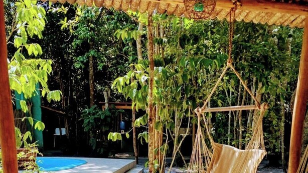 A hammock chair, a cushioned bench, and a small table with a vase in a shaded outdoor area with a pool in the background. - Chalés Guigó (Prado)