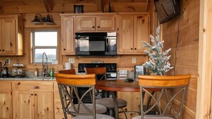 A wooden cabin interior with a kitchen area featuring wooden cabinets, a microwave, and a small Christmas tree. There are two wooden bar stools and a dining table.