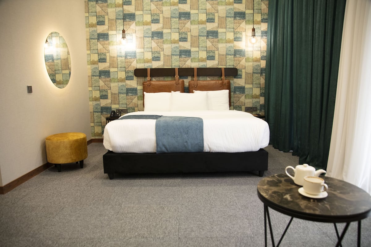 Een net opgemaakt bed met hoofdbord, een klein rond voetenbankje, een tafel met theepot en kopje, en een behang met patroon.