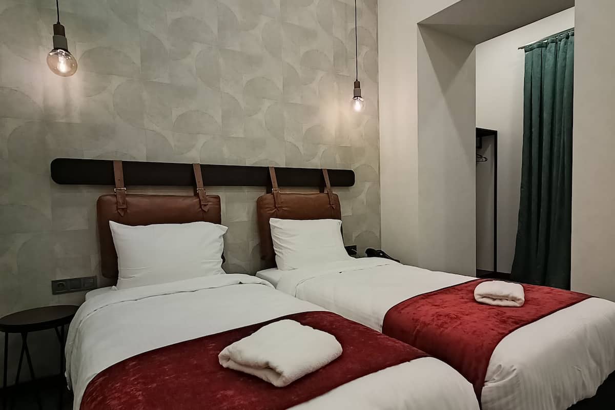 Een hotelkamer met twee bedden, elk met een rood-witte spreidek en een hoofdbord met bruine leren banden. Er hangen twee lampen en een klein tafeltje staat naast de bedden.