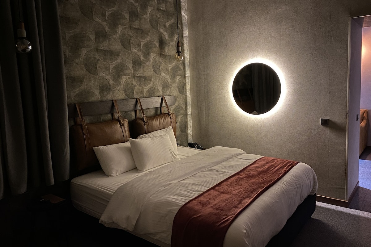Een keurig opgemaakt bed met een hoofdbord, een ronde spiegel en een wandlamp.