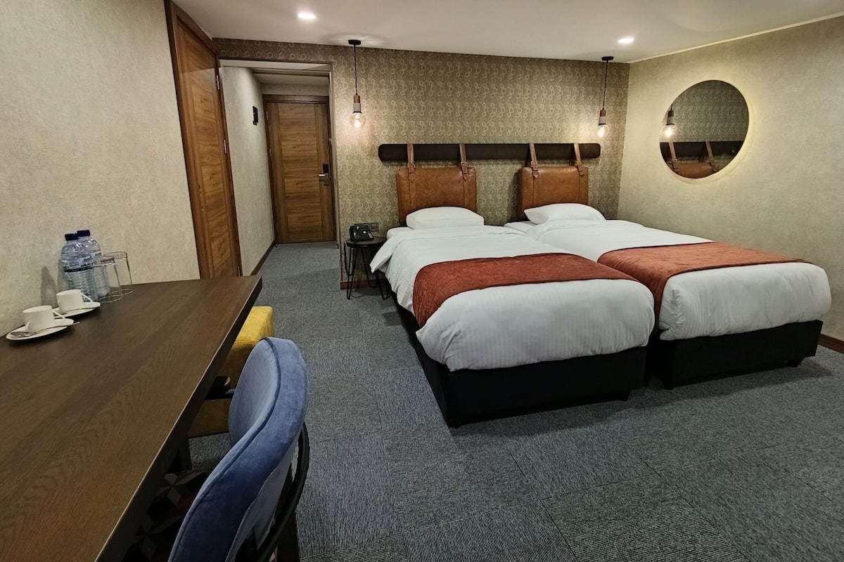 Een hotelkamer met twee bedden, een bureau met stoel, een klein tafeltje met bekers en een waterfles, en een spiegel aan de muur.