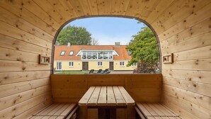 Un sauna en bois, avec des bancs et une table, une fenêtre en arc de cercle et une vue sur un bâtiment jaune avec un toit rouge.