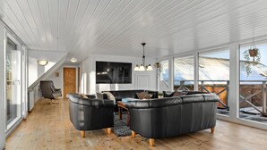 Un vaste espace de vie au design moderne, comprenant un ensemble de canapés en cuir noir, une table basse en bois et un grand téléviseur à écran plat fixé au mur.