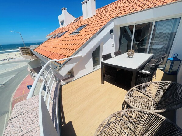 A balcony with a table and chairs, a view of the beach, and a tiled roof.
