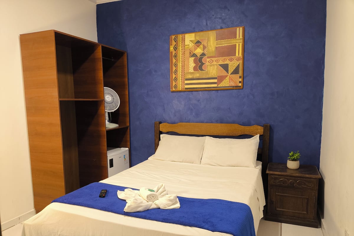 A neatly made bed with a blue towel, a wooden wardrobe, a small bedside table with a plant, and a wall art piece.