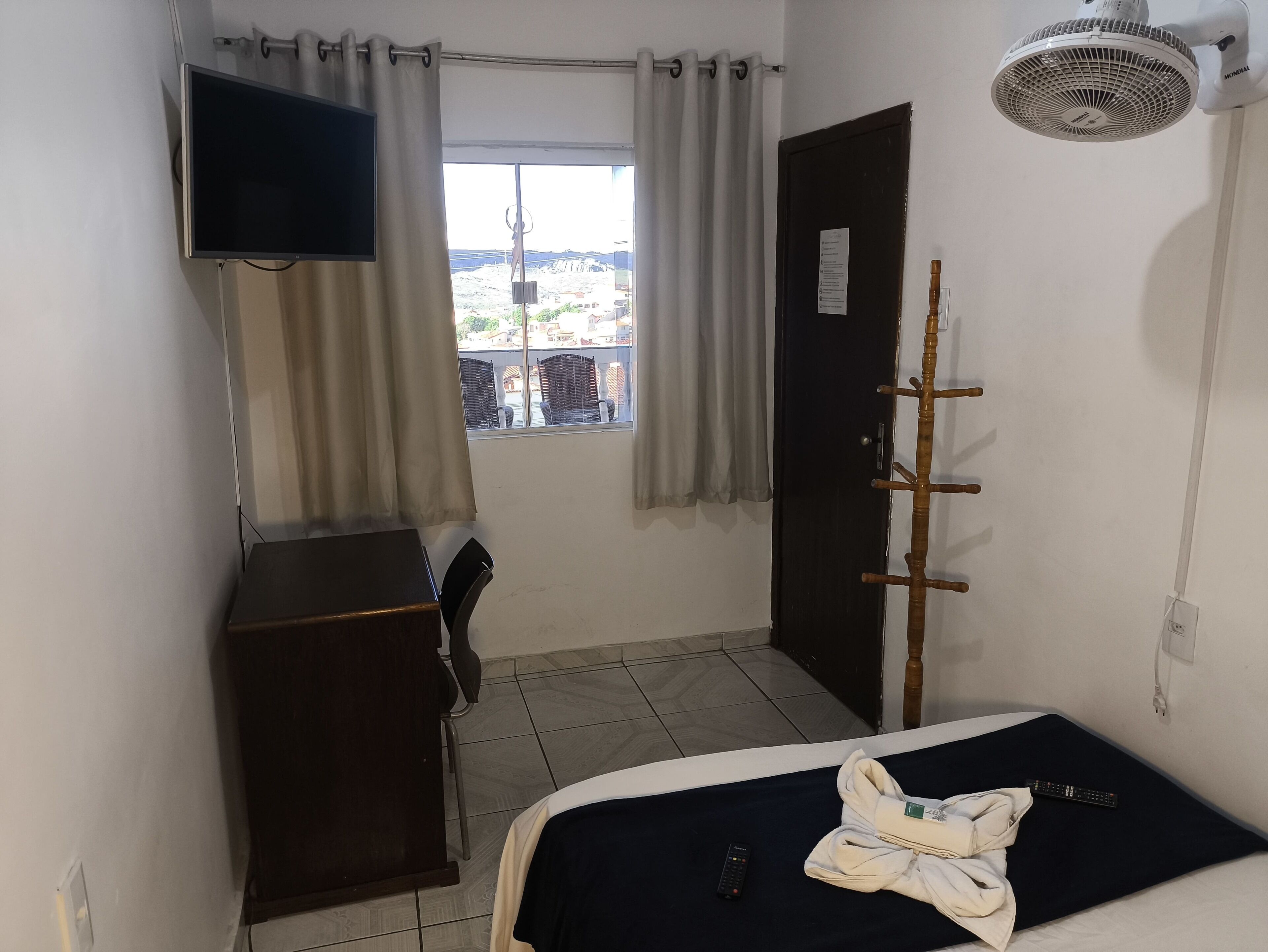 Ein Zimmer mit einem Bett, einem an der Wand befestigten Fernseher, einem Fenster mit Vorhängen, einem Kleiderständer und einer Tür.
