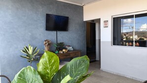 A modern living room with a flat-screen TV mounted on the wall, a wooden side table with books and decorative items, and large green plants in the foreground. - HOTEL VILLA REAL (Diamantina)