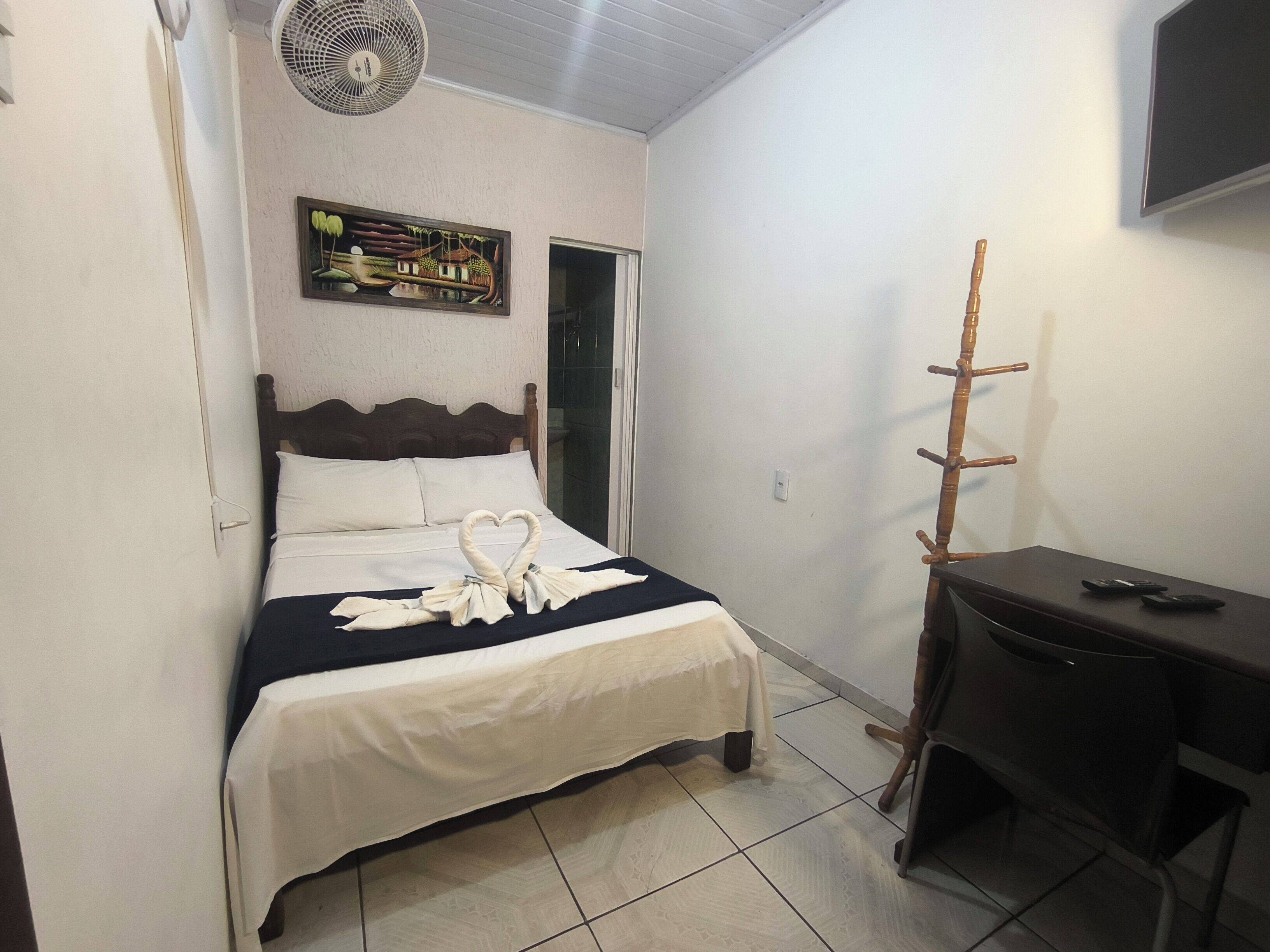 Une chambre d’hôtel avec un lit, un bureau, une chaise, un ventilateur, un tableau au mur et un téléviseur.