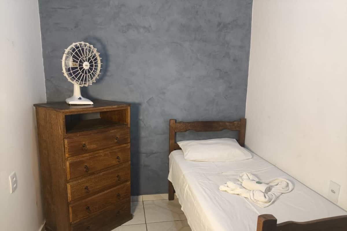 A bedroom with a wooden dresser, a bed with white bedding, and a white fan on the dresser.