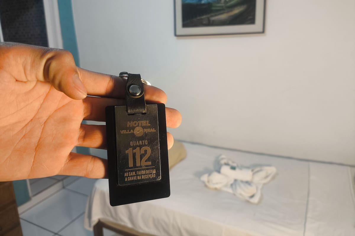 A hand holding a keycard for Hotel Villa Real, room number 112, with a message in Portuguese.