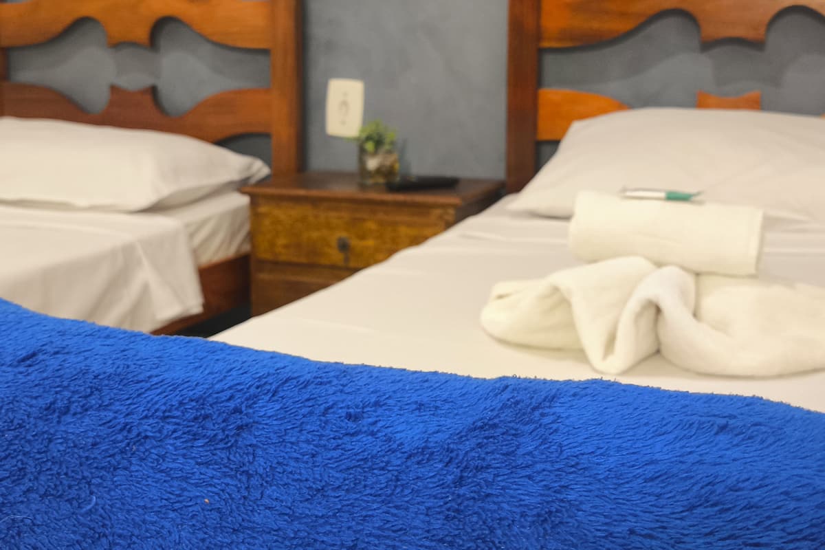 A hotel room with two beds, wooden headboards, white linens, a blue throw blanket, and a nightstand with a lamp and a small vase.