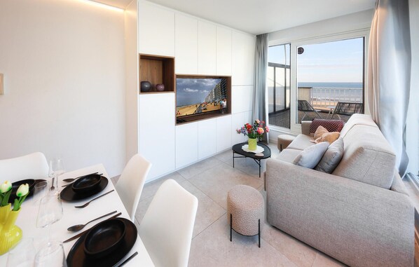 A modern living room with a sofa, dining table, and a view of the beach through a large window.