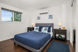 A bedroom with a bed, two bedside tables, a nightstand, an air conditioning unit, and artwork on the wall.