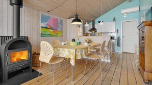 A cozy room with a wood-burning stove, a dining table with chairs, a kitchen area, and a large painting on the wall. - Amazing home in Broager with kitchen (Broager)