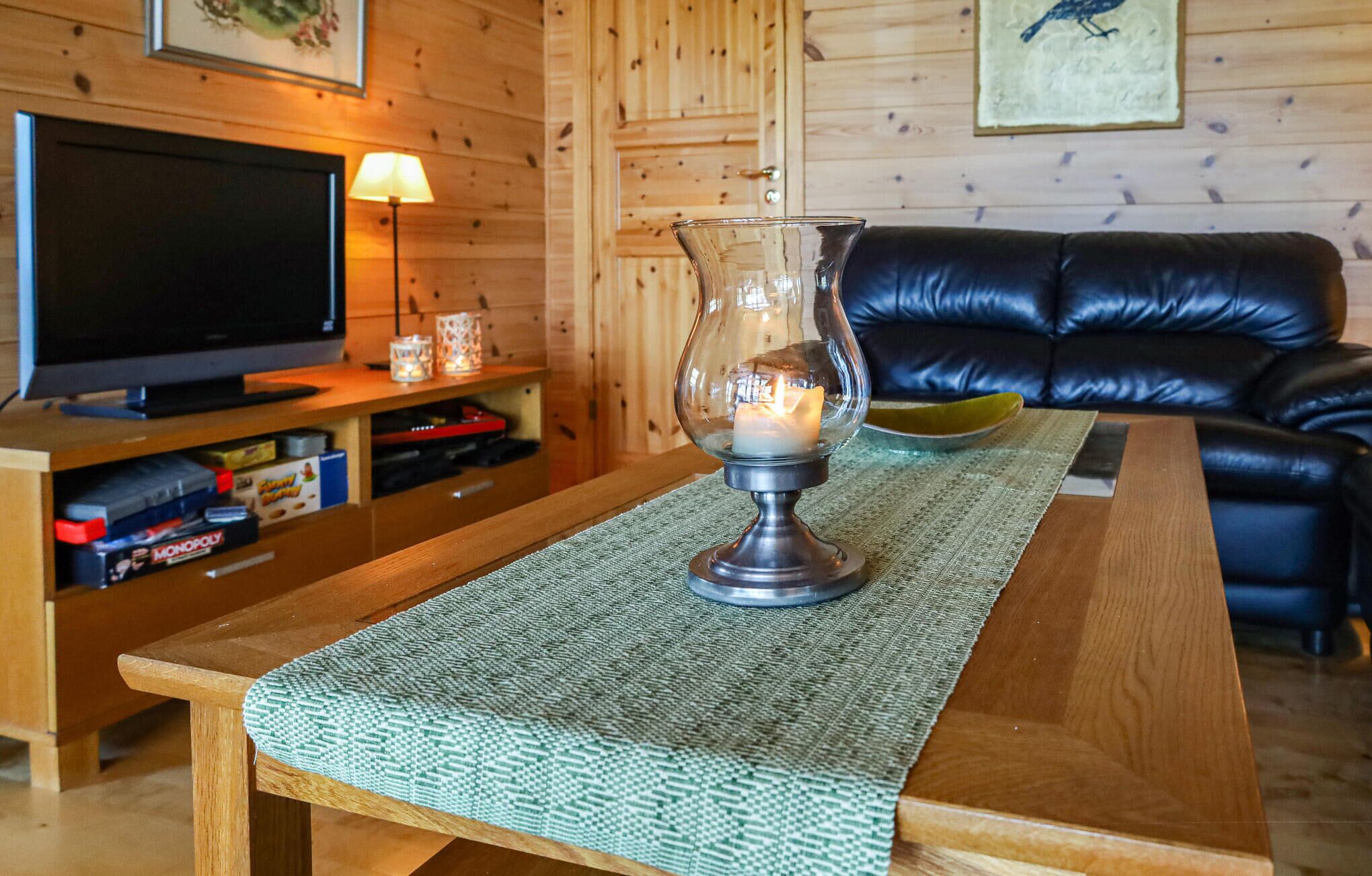 A wooden table with a woven placemat, a glass candle holder with a lit candle, a black leather sofa, a television on a wooden stand, and a framed picture on the wall.