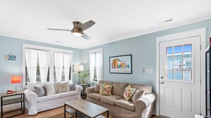 A living room with a beige sofa, a coffee table, and a ceiling fan. There is a door with a window, a lamp, and a framed picture on the wall. - Cheerful 3BR/2BA - just steps to the beach! (Oak Island)