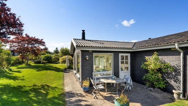 Une maison à l’extérieur sombre, avec une cheminée bien visible, une terrasse dallée avec une table et des chaises, et un jardin avec de la pelouse et des arbres.
