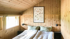 A wooden cabin room with a bed, two nightstands, a framed bird illustration on the wall, and a window with curtains.