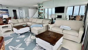 Ein Wohnzimmer mit einem Ecksofa, zwei Couchtischen, einem Teppich mit einem Seesternmuster, einer Treppe und einem großen Fenster mit Blick auf die Stadt.