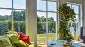 A room with a large window, a green sofa with red cushions, a round table with a tray, and a hanging plant. - 4 bedroom stunning home in Ramdala (Ramdala)