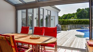 A sunlit patio with a wooden table and red chairs, overlooking a swimming pool and lush greenery. - 4 bedroom stunning home in Ramdala (Ramdala)