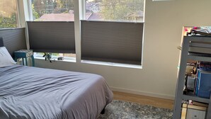 A bedroom with a bed, two windows with blinds, a small table with a plant, and a bookshelf with books and items. - Modern, spacious four-bedroom home on quiet street (Seattle)