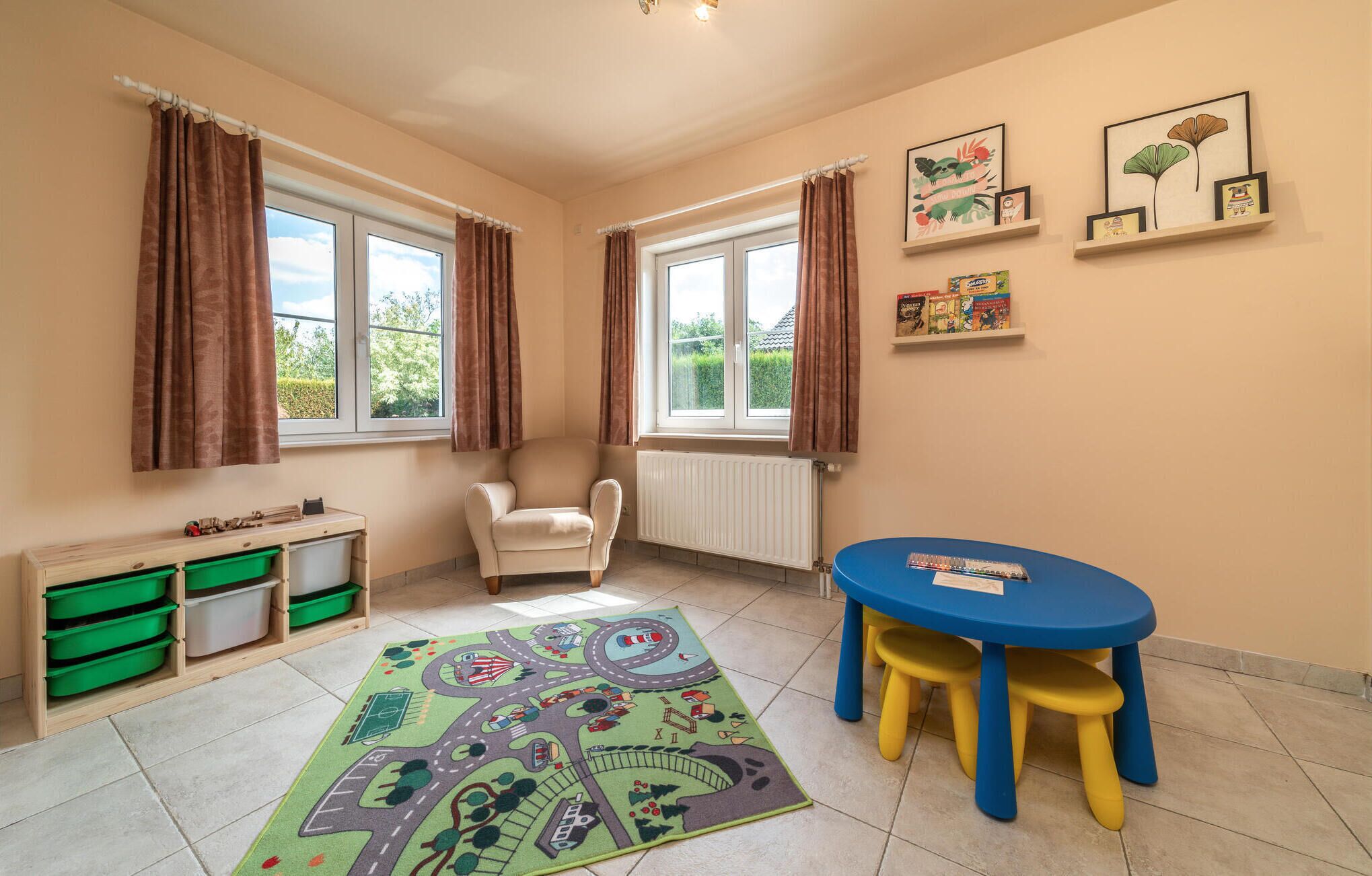 Ein Zimmer mit zwei Fenstern, einem Heizkörper, einem grünen Schrank, einem weißen Stuhl, einem blauen Tisch mit gelben Hocker und einem bunten Teppich.