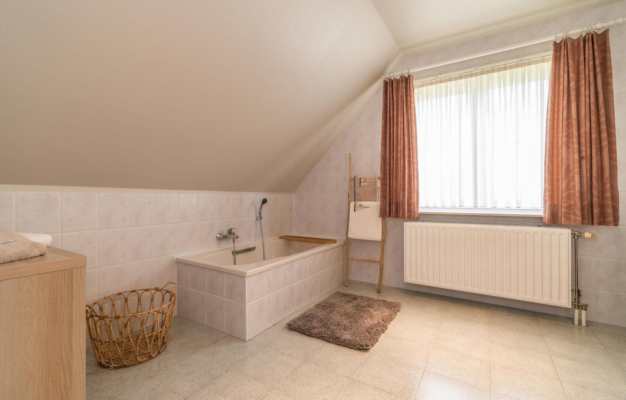 Un baño con bañera, un mueble de madera, una cesta de mimbre, un radiador y una ventana con cortinas.