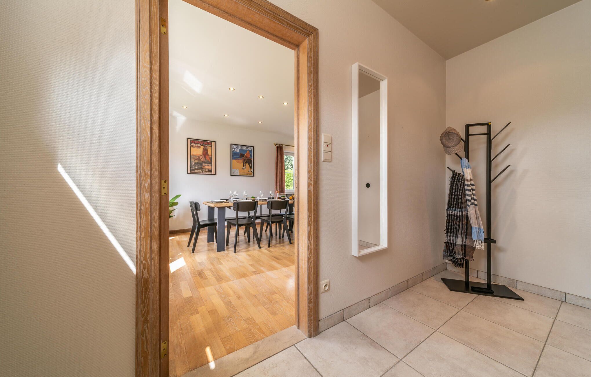 Um corredor amplo com porta de madeira que dá para uma sala de jantar. Há um porta-chapéus com chapéus e cachecóis, e um interruptor de luz fixado na parede.