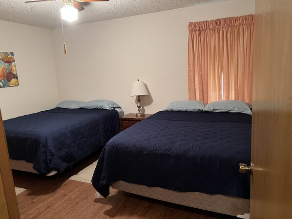 Una habitación con dos camas, una mesita de noche con lámpara, un ventilador de techo y un cuadro en la pared.