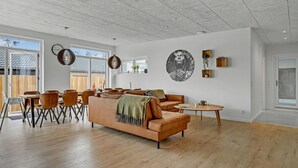 A spacious living area with a large sofa, dining table, and chairs. There are pendant lights, a fireplace, and a large window providing natural light.