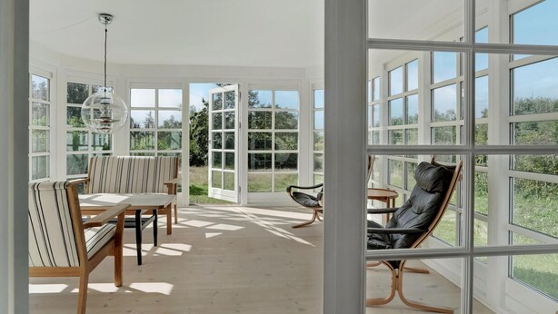 Un espacio habitable amplio y luminoso con comedor, zona de estar con una butaca de cuero negro y ventanas grandes con vistas al exterior.