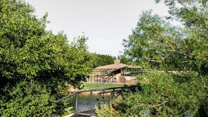 Un chalet rustique doté d’une terrasse en bois et d’un petit pont enjambant un étang, le tout entouré d’une végétation luxuriante.