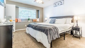 Une chambre à coucher comprenant un lit, une table de chevet, une commode, un ventilateur de plafond, une fenêtre et une œuvre d’art accrochée au mur.