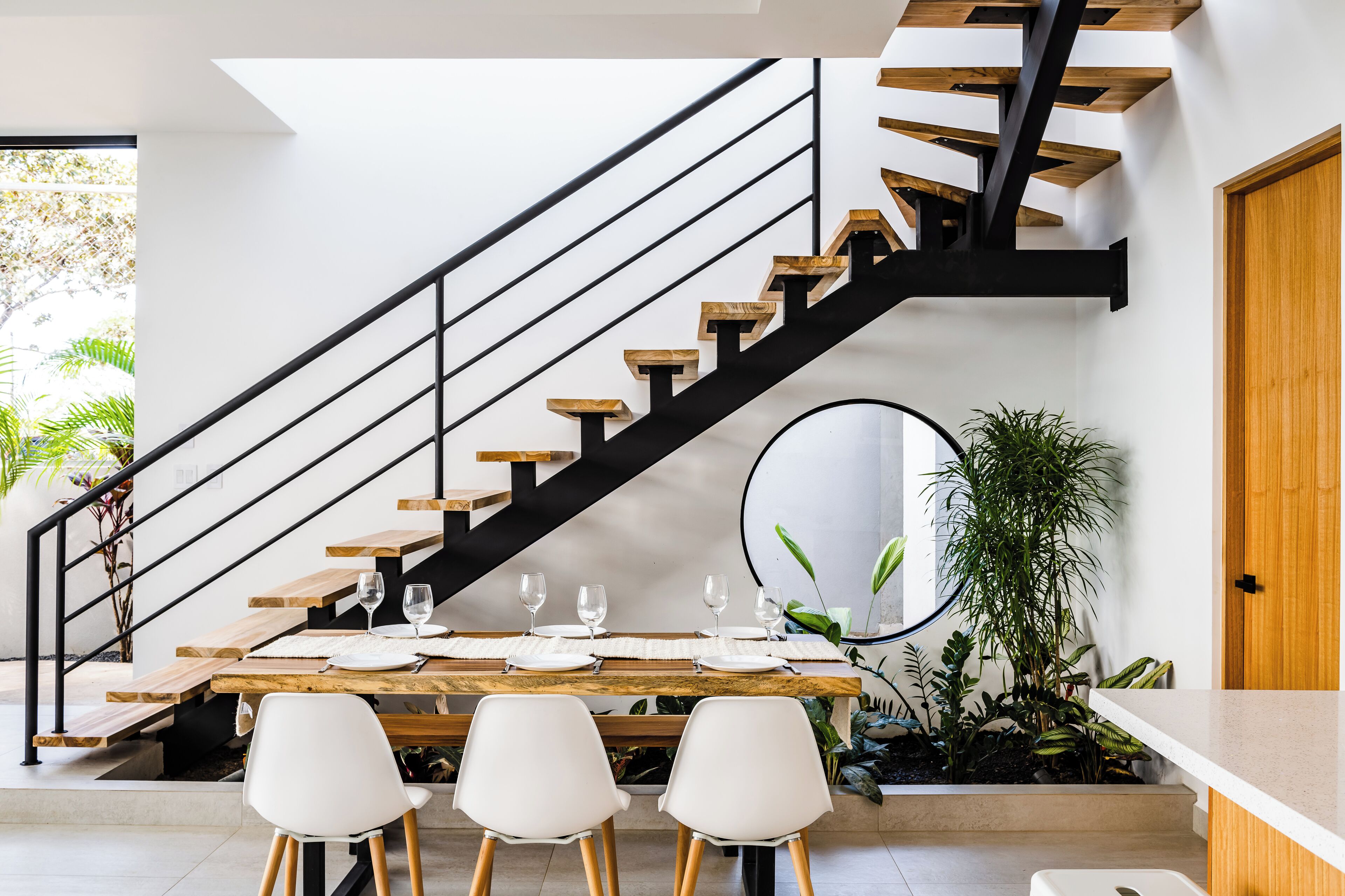 A modern dining area with a wooden table set for four, a black staircase with wooden steps, and a large mirror on the wall.