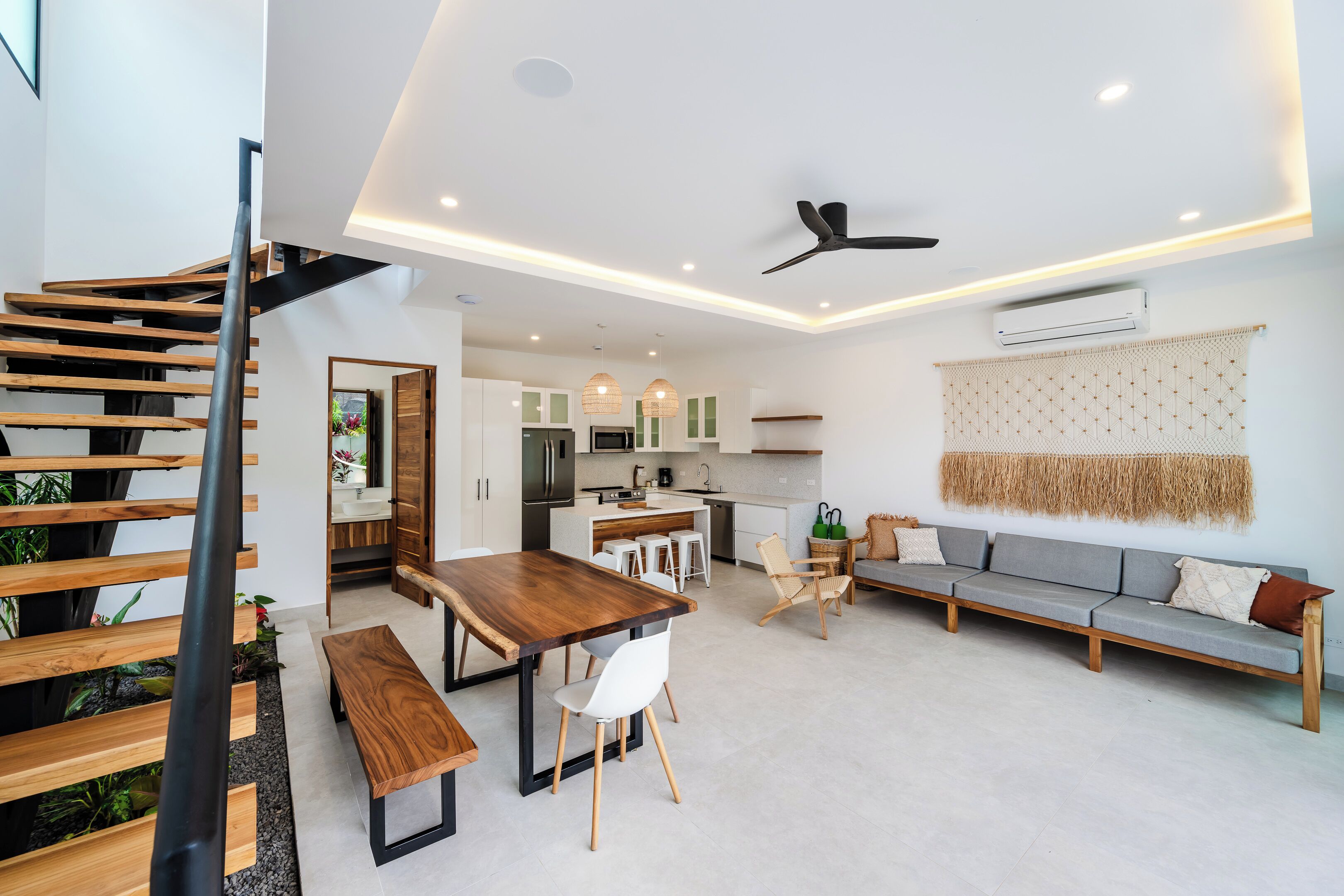A modern kitchen with white cabinets, a dining area with a wooden table and chairs, a gray sofa, and a staircase with wooden steps.