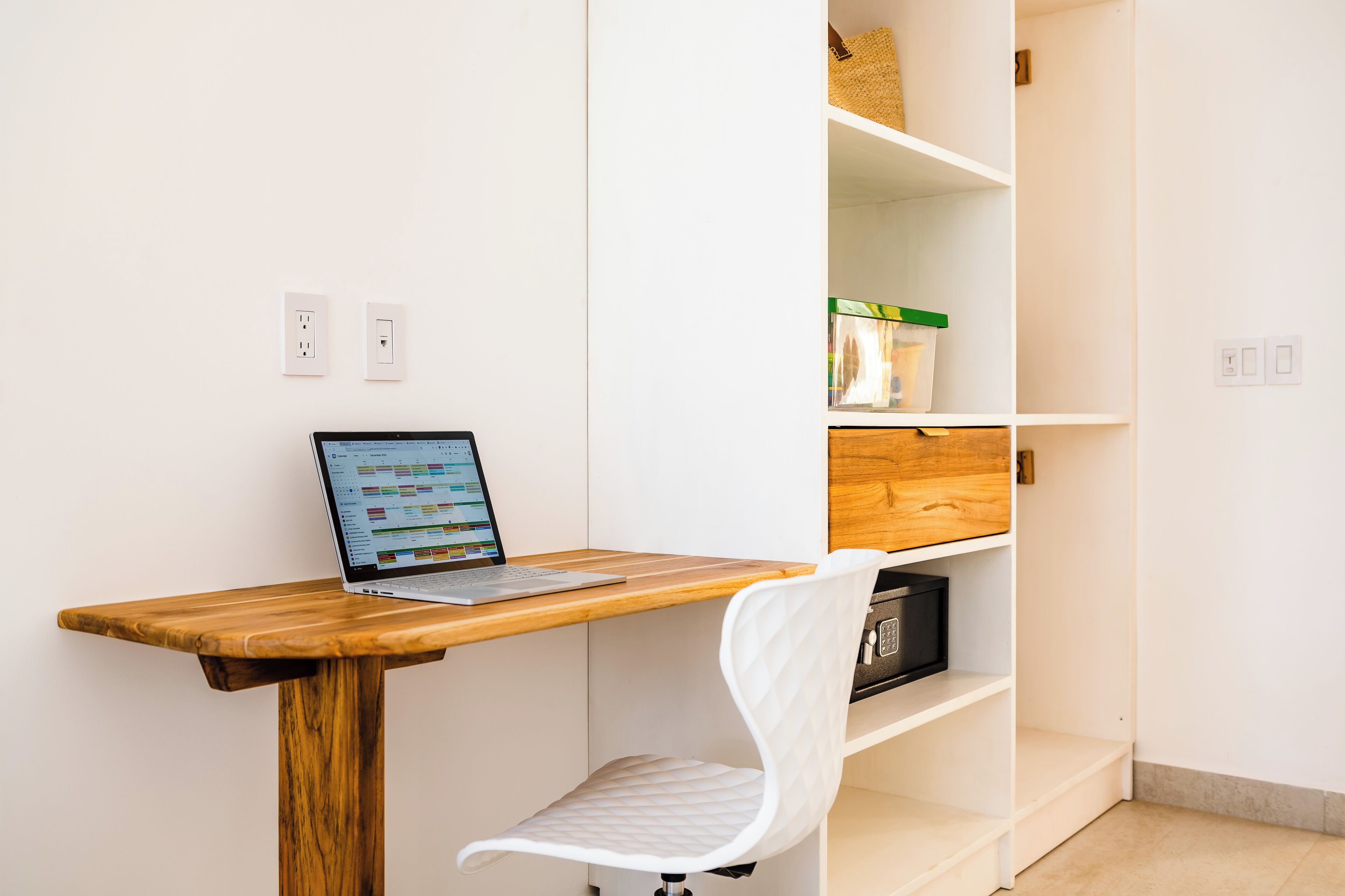 Un espacio de trabajo con una computadora portátil sobre un escritorio de madera, una silla blanca y un estante blanco con cajas de almacenamiento.