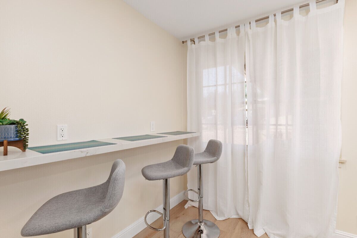 A room with a white wall, a window with white curtains, and two grey bar stools.