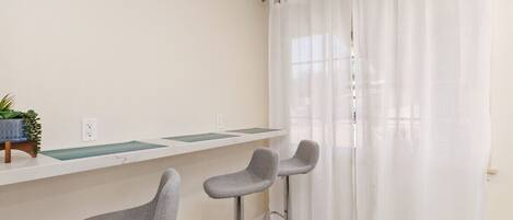 A room with a white wall, a window with white curtains, and two grey bar stools.