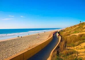Ein Strand mit einer gebogenen Betonbarriere, die Sand und Grünfläche voneinander trennt.