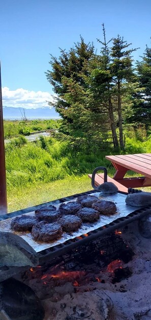 A grill with several burgers cooking outdoors, surrounded by a grassy landscape and trees.