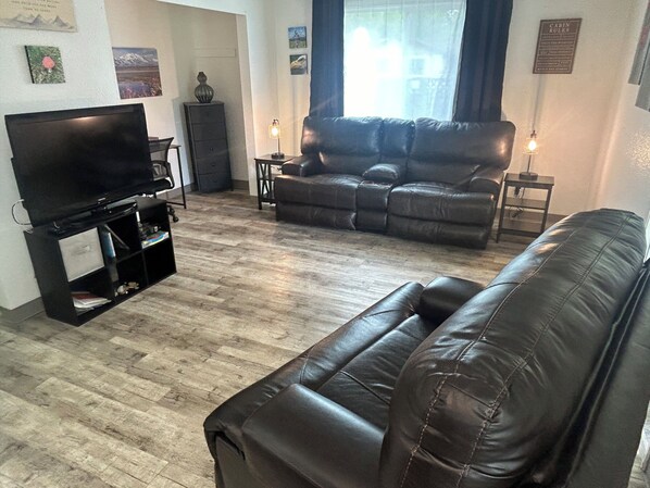 A living room with a black leather sofa, a flat-screen TV on a stand, and a wooden floor.