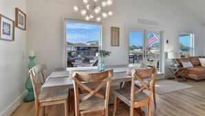 A spacious living room with a large window, a wooden dining table with chairs, a chandelier, and a view of the beach.
